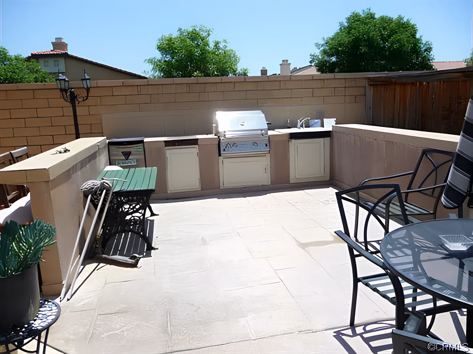 Outdoor patio area with a built-in barbecue grill, sink, and storage cabinets along a brick wall. There is a green table with a bench on the left side and a glass-top table with metal chairs on the right side. Trees and rooftops are visible in the background under a clear blue sky.