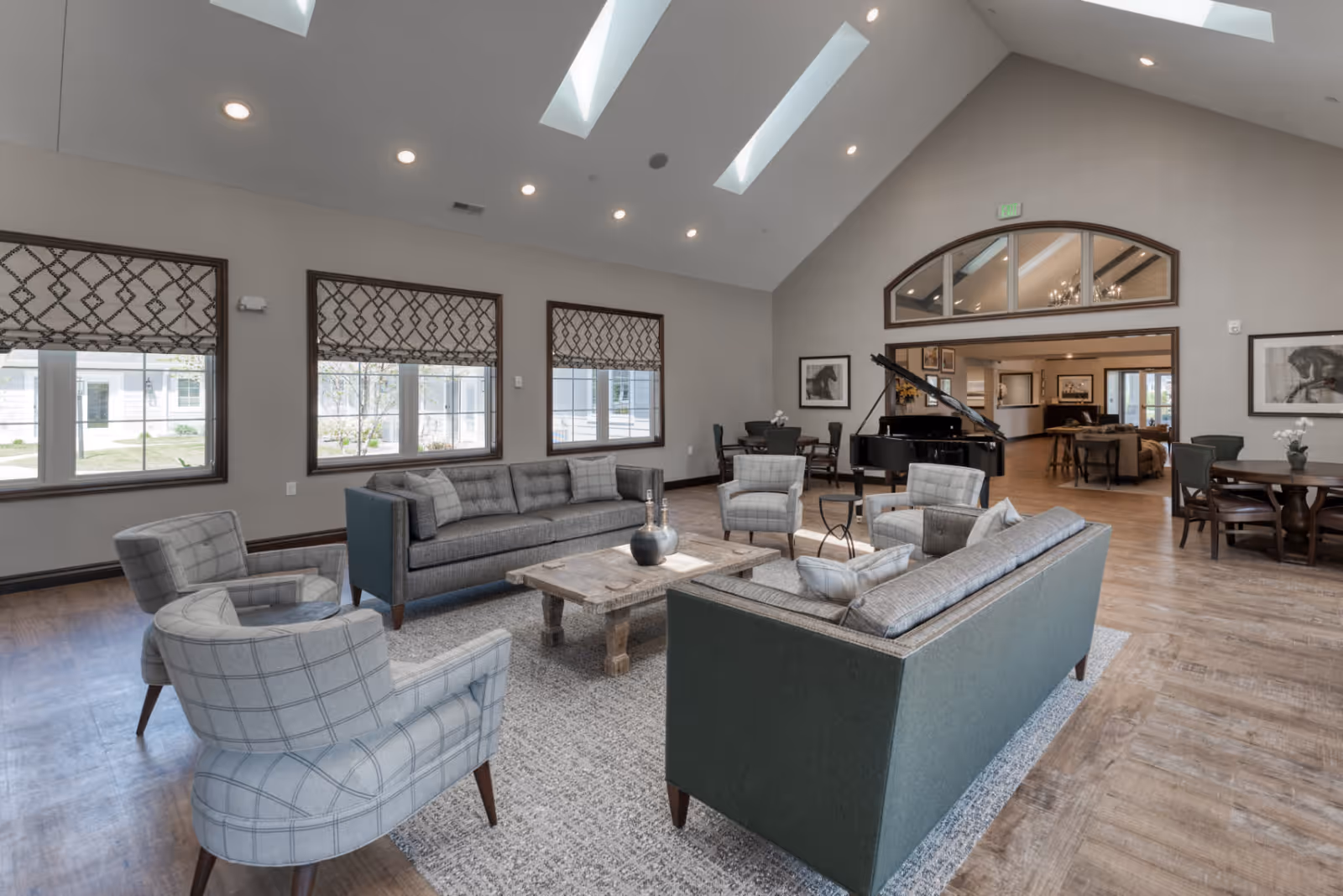 A spacious senior living facility common area with high vaulted ceilings and skylights. The room features two gray sofas, four plaid armchairs arranged around a wooden coffee table on a large area rug. There are three windows with patterned Roman shades letting in natural light. In the background, a black grand piano is visible near a round table with chairs, and an open doorway leads to another room with more seating and artwork on the walls.