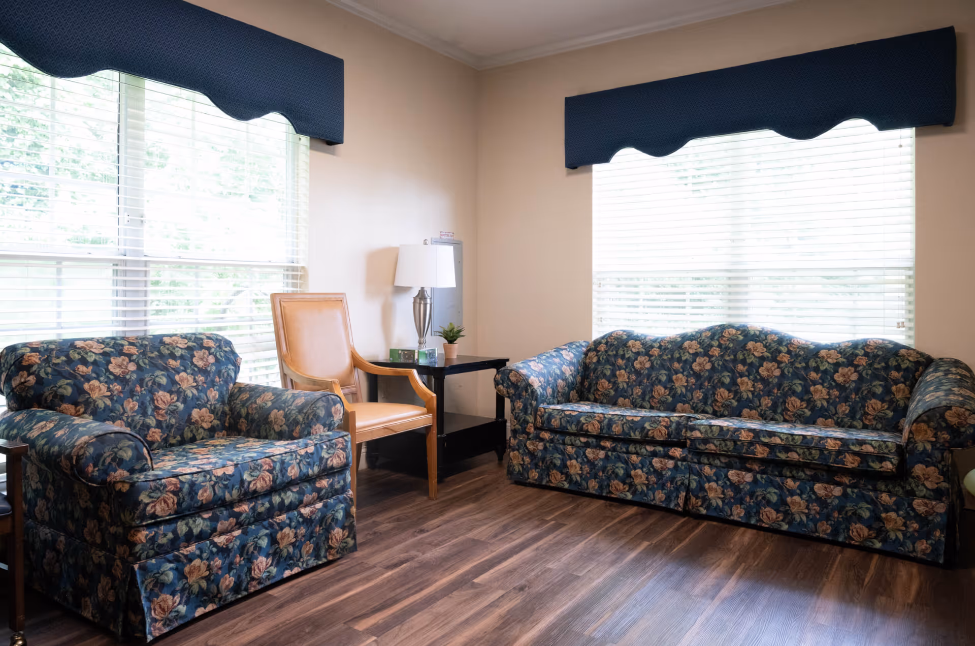 Sunlit sitting room with a floral-patterned sofa and matching armchair, wooden floor, large windows with blinds and blue valances, and a side table with a lamp.