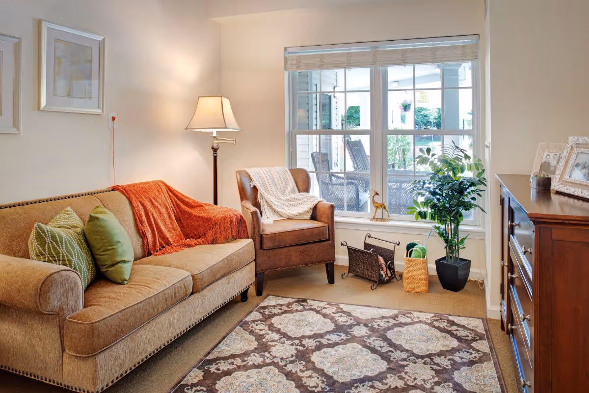A cozy living room with a beige sofa adorned with green pillows and an orange throw blanket, a brown armchair with a white knitted throw, a floor lamp, a patterned area rug, a wooden dresser with framed photos, a potted plant, and a large window showing an outdoor patio with chairs.