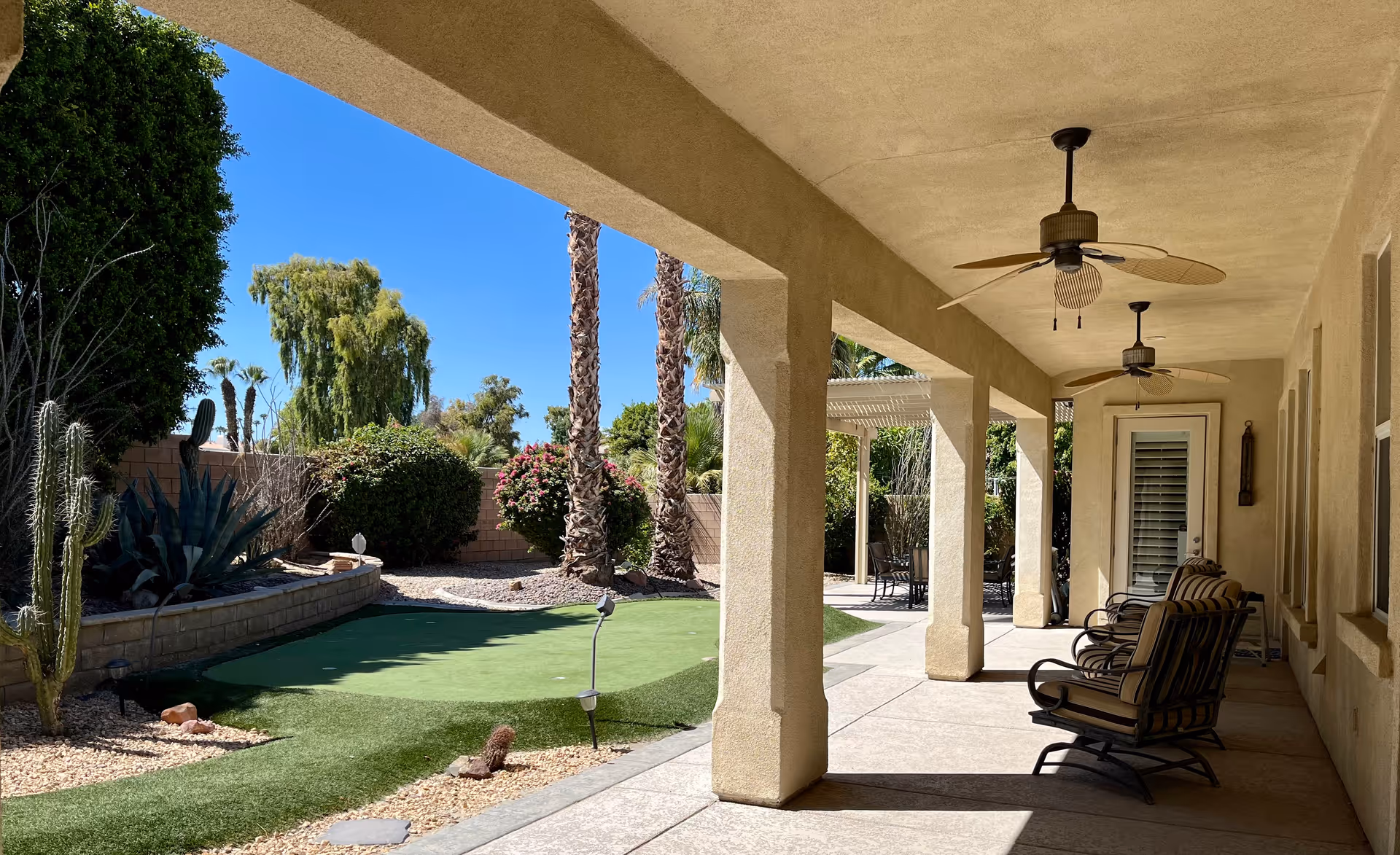 A covered outdoor patio area with ceiling fans and cushioned chairs overlooking a landscaped garden with palm trees, bushes, and a small putting green under a clear blue sky.