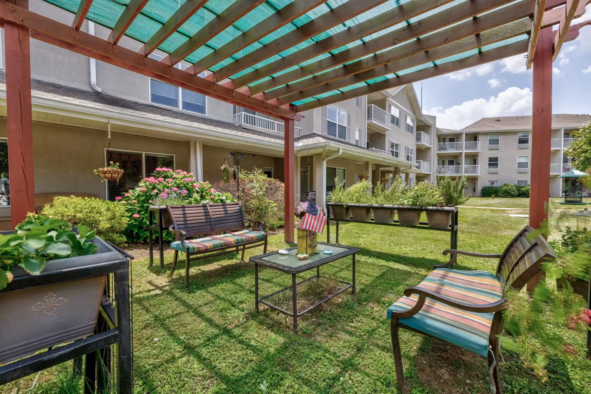 Courtyard seating area under a pergola with benches, a coffee table, and potted plants in front of a multi-story senior living building.