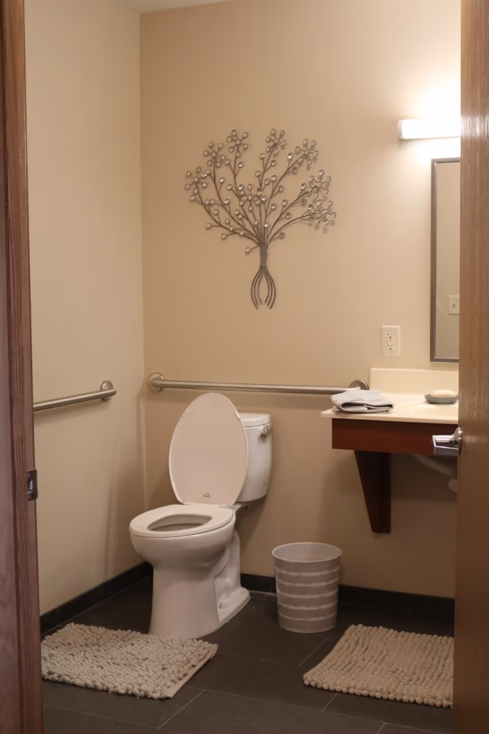 A clean bathroom with a white toilet, two beige textured rugs on the dark floor, a beige wall with a decorative metal wall art shaped like a tree, a small trash bin, and a wooden vanity with a white countertop holding folded towels and a soap dish. There are grab bars on the walls near the toilet.