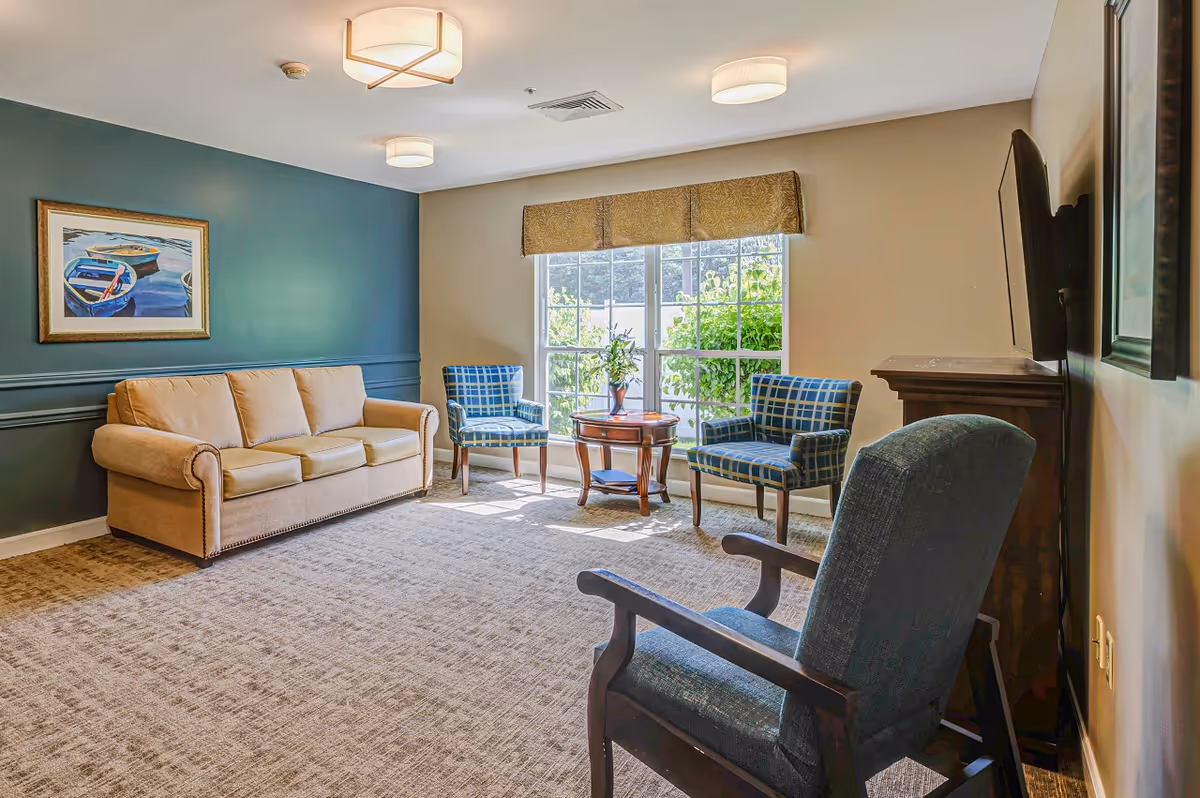 Sunlit seating area with a beige sofa, plaid armchairs, a small table, and a wall-mounted TV next to a large window.