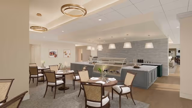 A modern dining area in a senior living facility featuring round tables set with plates and glasses, surrounded by cushioned wooden chairs. The room includes a serving counter with a glass display case and pendant lights hanging above. The decor is neutral with light-colored walls, carpeted and wooden flooring, and framed artwork on the walls.