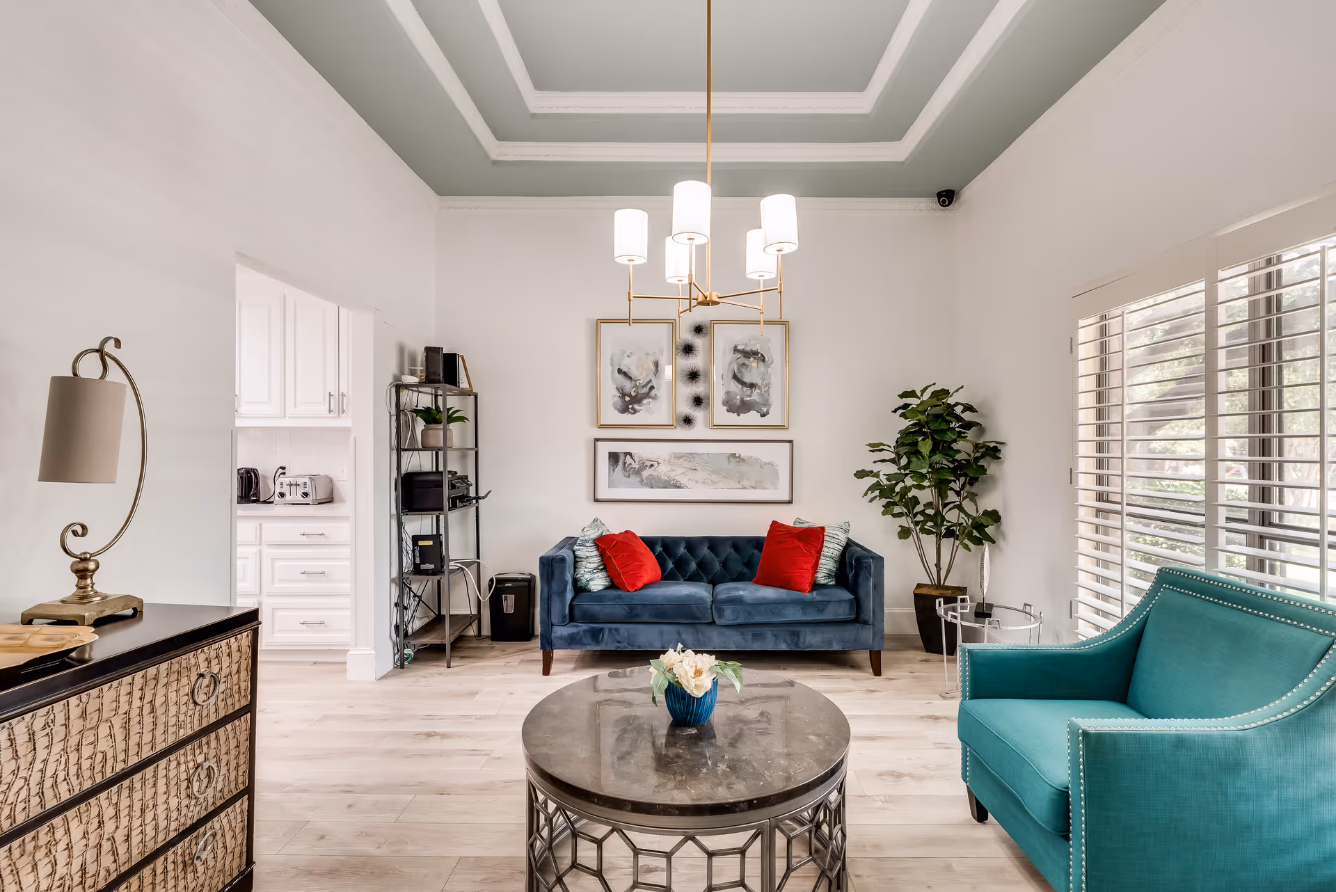 A bright and modern living room with a blue velvet sofa adorned with red and patterned pillows, a teal armchair, a round coffee table with a small flower arrangement, a tall potted plant, and a decorative lamp on a textured cabinet. The room features a tray ceiling with a chandelier, light wood flooring, and large windows with white blinds letting in natural light. A doorway leads to a white kitchen area with visible appliances.
