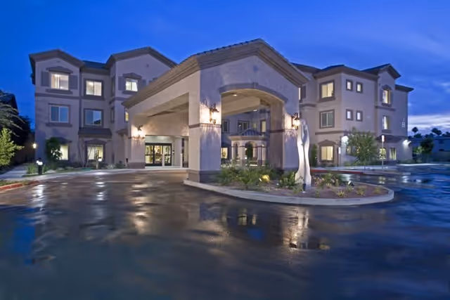 Exterior view of a three-story senior living facility building at dusk with lights on and a covered entrance with columns. The driveway and surrounding landscaping are visible with a modern sculpture near the entrance.