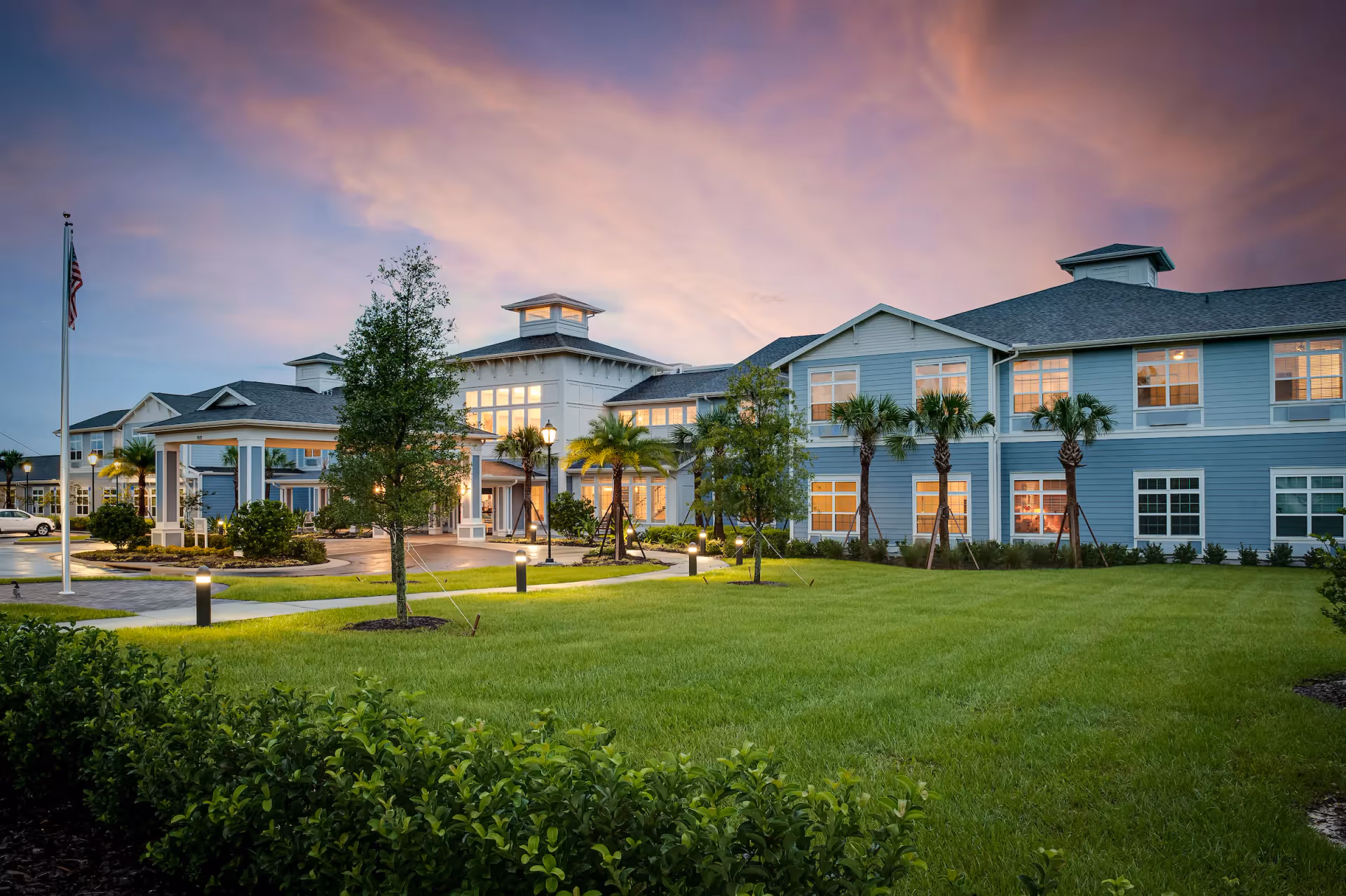Exterior view of Sancerre At Orange City senior living facility at dusk, showing a large two-story building with blue siding, multiple windows lit from inside, palm trees, a well-maintained lawn, and a flagpole with an American flag.