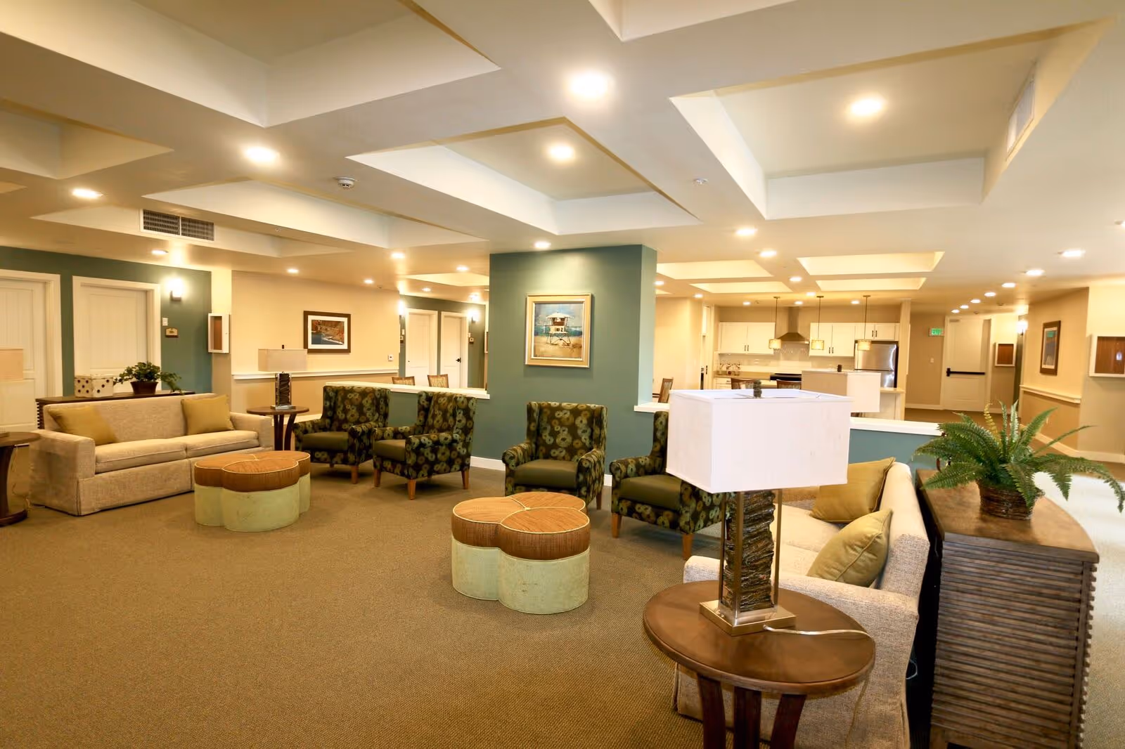 A spacious and well-lit living room area in a senior living facility with beige sofas, patterned armchairs, round ottomans, a wooden side table with a lamp, and a potted plant. The room has a coffered ceiling with recessed lighting and a kitchen area visible in the background.