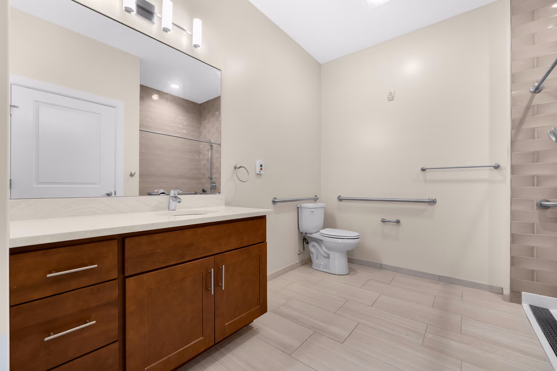 Spacious accessible bathroom with a wooden vanity and sink, large mirror, toilet, multiple grab bars, and a tiled shower.