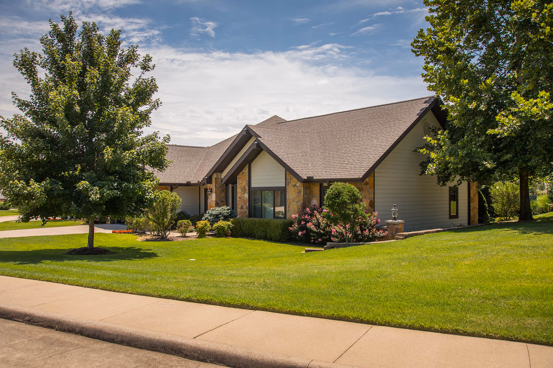 Single-story residential building with stone accents, a manicured lawn, flowering shrubs and trees bordered by a sidewalk under a partly cloudy sky.