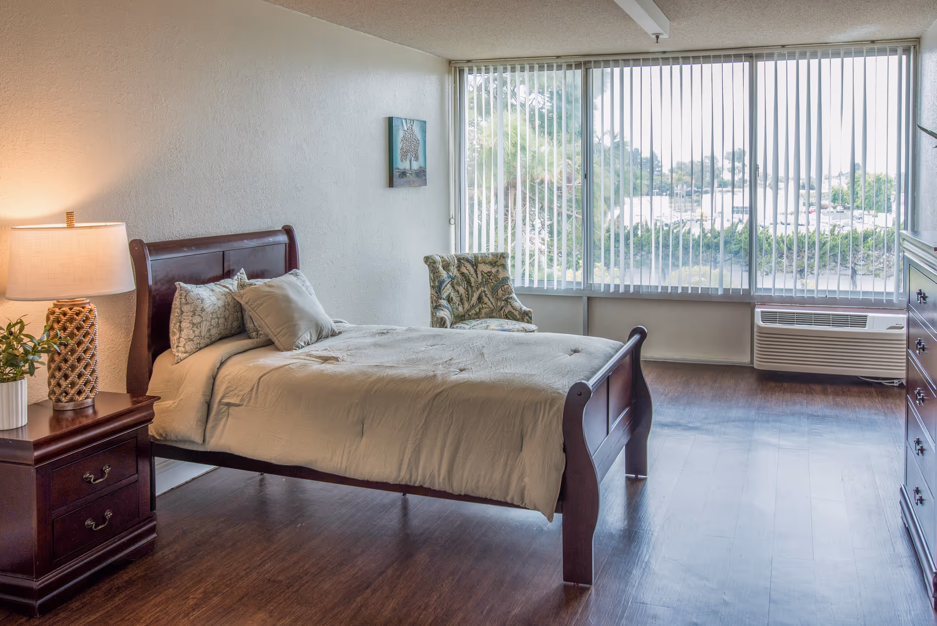 A bedroom with a wooden bed frame and beige bedding, a wooden nightstand with a lamp and a small plant, a patterned armchair near a large window with vertical blinds, and a wooden dresser. The room has wooden flooring and a view of greenery and buildings outside the window.