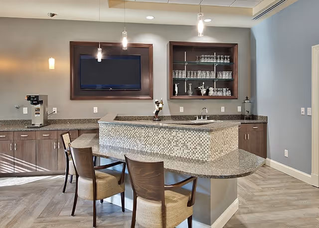 A modern kitchen area in a senior living facility featuring a granite countertop island with mosaic tile front, two wooden chairs with cushioned seats, a mounted flat-screen TV, glass shelves with various glassware, a coffee machine on the counter, and pendant lights hanging from the ceiling.