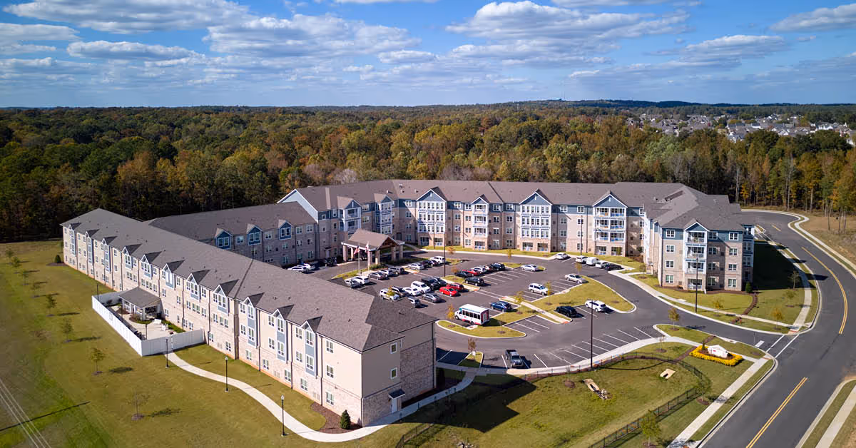 Aerial view of a large senior living facility named The Crossings at North River, showing a multi-story building with a parking lot filled with cars in the center, surrounded by green lawns and trees under a partly cloudy sky.