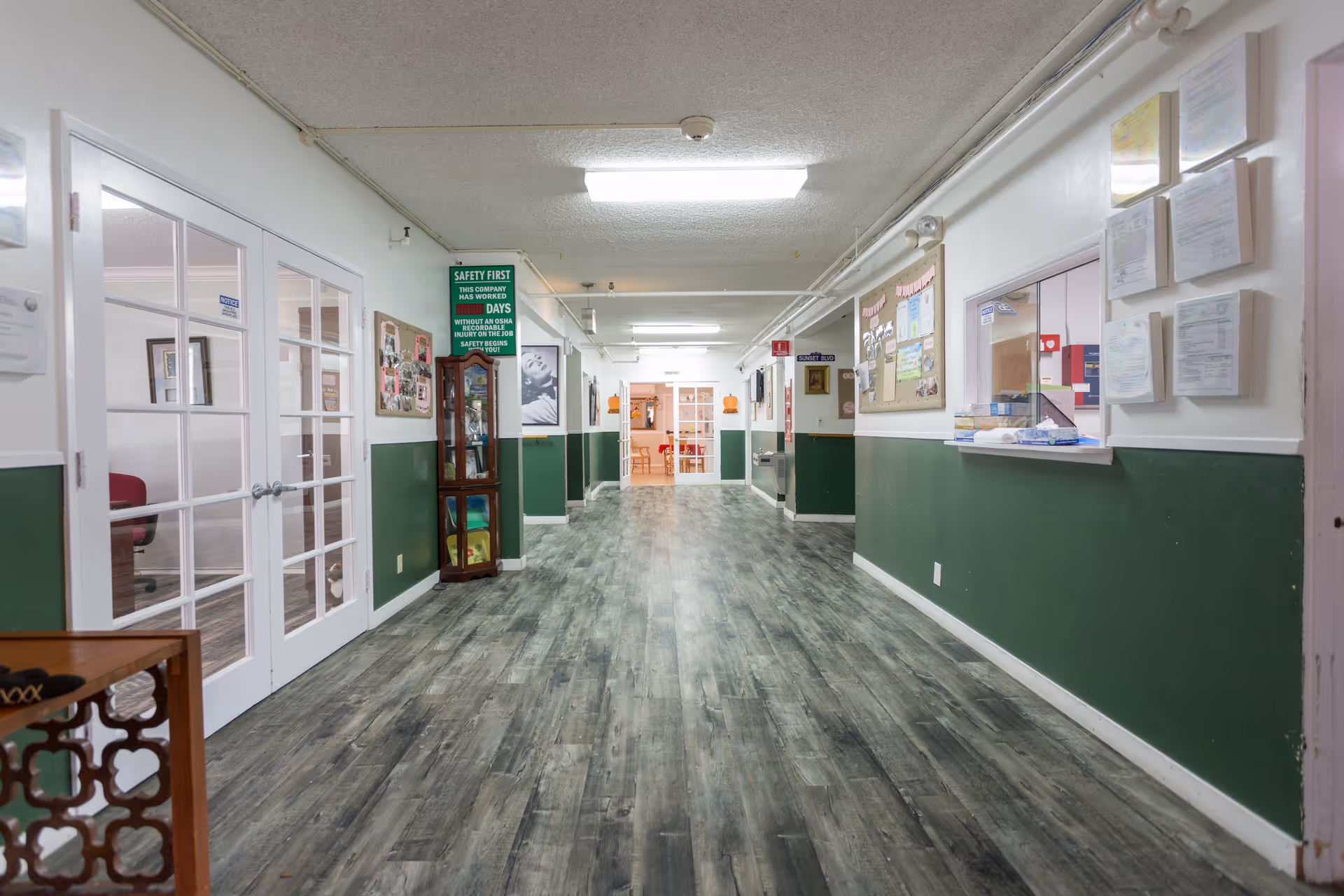Long interior hallway with green half-walls, wood-look flooring, bulletin boards and a reception window.