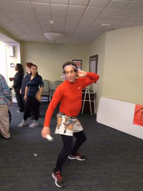 A man in a red long-sleeve shirt, black pants, and a colorful apron is posing playfully in a room with several other people in the background. The room has light-colored walls, a carpeted floor, and some chairs and tables along the walls.