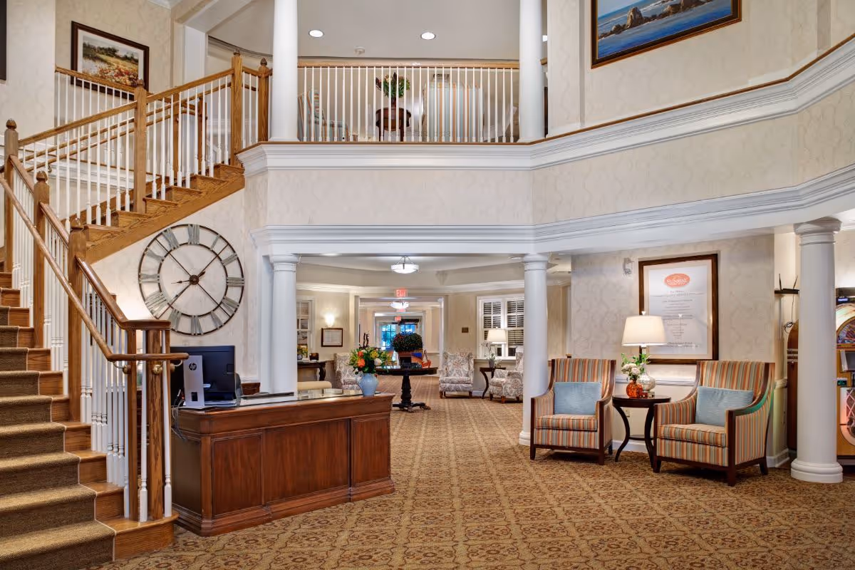 Lobby of a senior living facility with a wooden staircase, reception desk, seating area, and decorative columns.