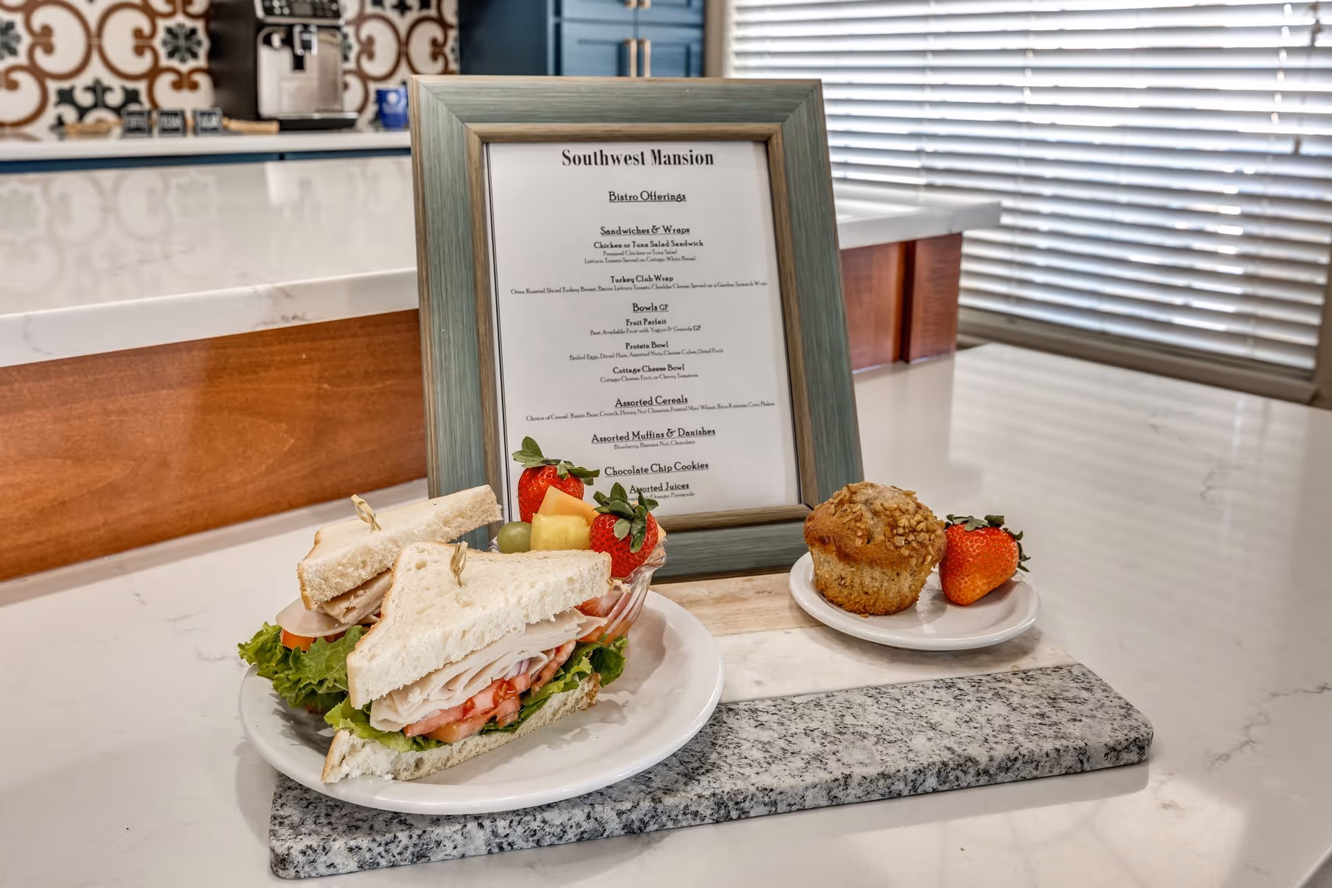 A plate with a turkey sandwich, lettuce, tomato, and grapes, alongside a small plate with a muffin and a strawberry, placed on a kitchen counter. Behind the food is a framed menu titled 'Southwest Mansion Bistro Offerings' with various food options listed. The background shows a kitchen area with a coffee machine and decorative tiles.
