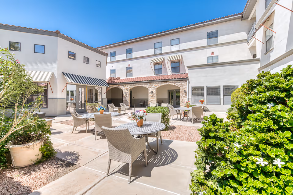 Outdoor courtyard area at The Woodmark at Sun City featuring several round tables with wicker chairs, potted plants with flowers on the tables, surrounded by white buildings with awnings and windows under a clear blue sky.