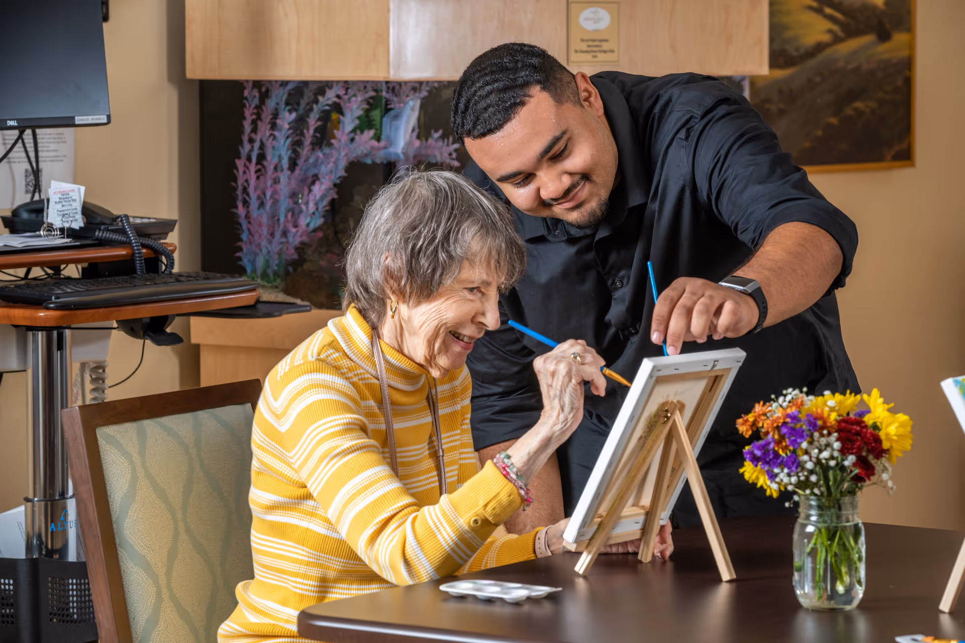An elderly woman in a yellow striped sweater is painting on a small canvas with a paintbrush, while a young man in a black shirt leans over and assists her. They are seated at a table with a vase of colorful flowers and painting supplies. In the background, there is a computer monitor and an aquarium.