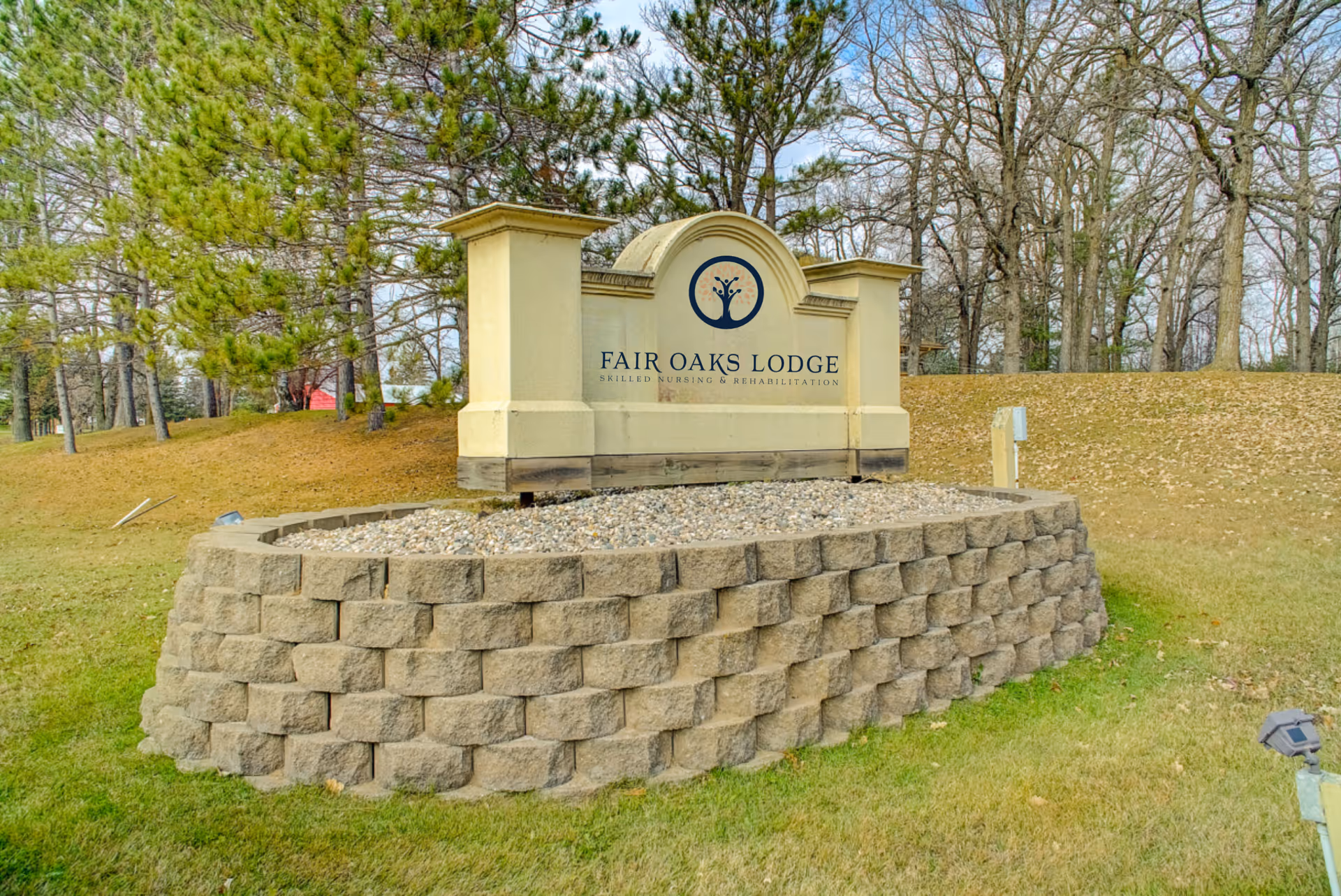 Outdoor stone sign for Fair Oaks Lodge Skilled Nursing & Rehabilitation surrounded by grass and trees in the background.