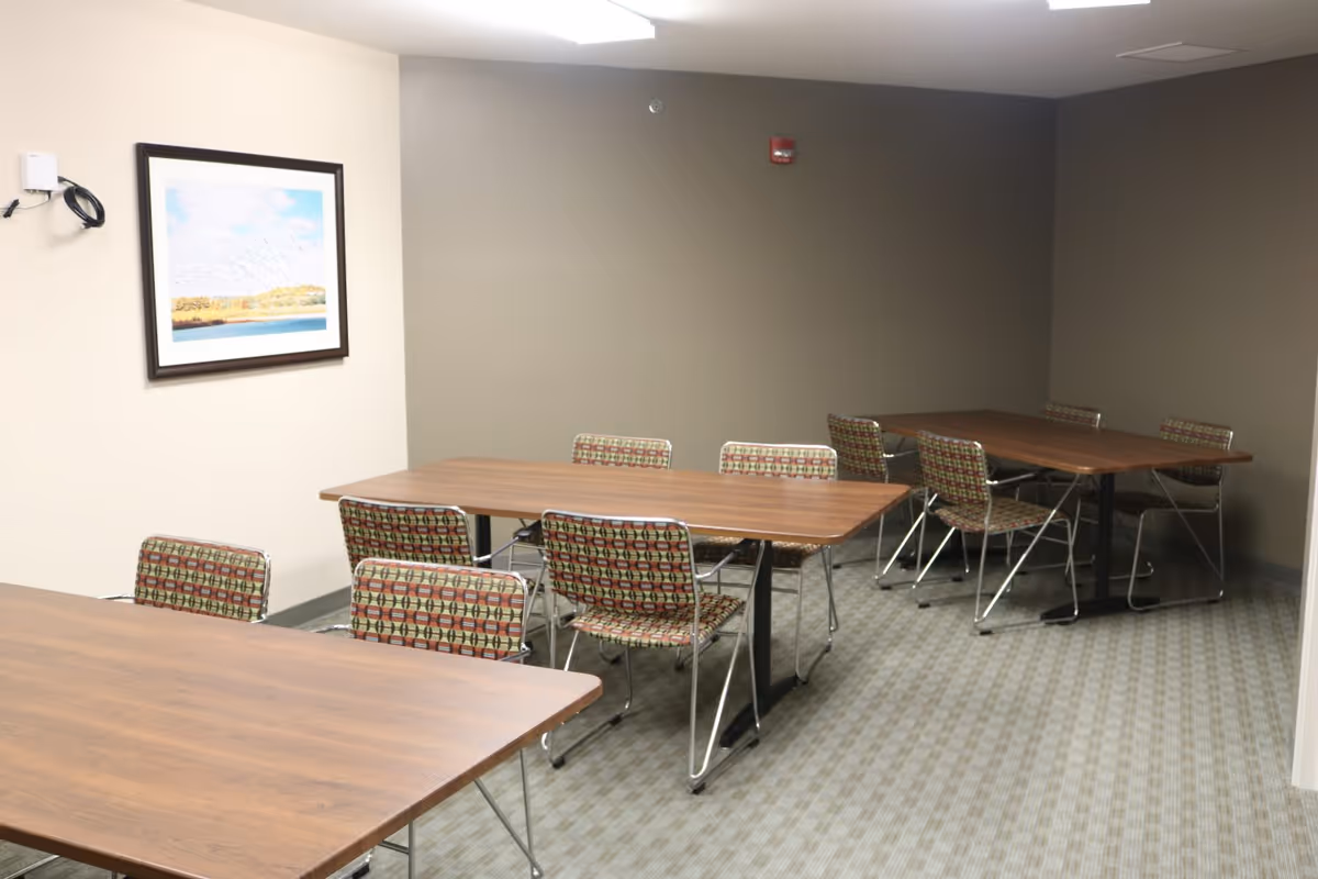 A room with three wooden tables and several patterned chairs arranged around them. The walls are painted in beige and gray tones, and there is a framed landscape picture hanging on one wall. The floor is carpeted with a subtle checkered pattern.