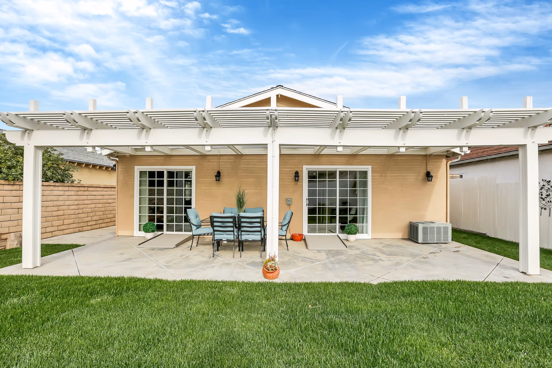 Outdoor patio area with a white pergola covering a concrete floor. There is a black metal table with six chairs that have light blue cushions. Two sliding glass doors lead into the building, which has a beige exterior wall. Green grass surrounds the patio, and there are potted plants and an air conditioning unit visible.