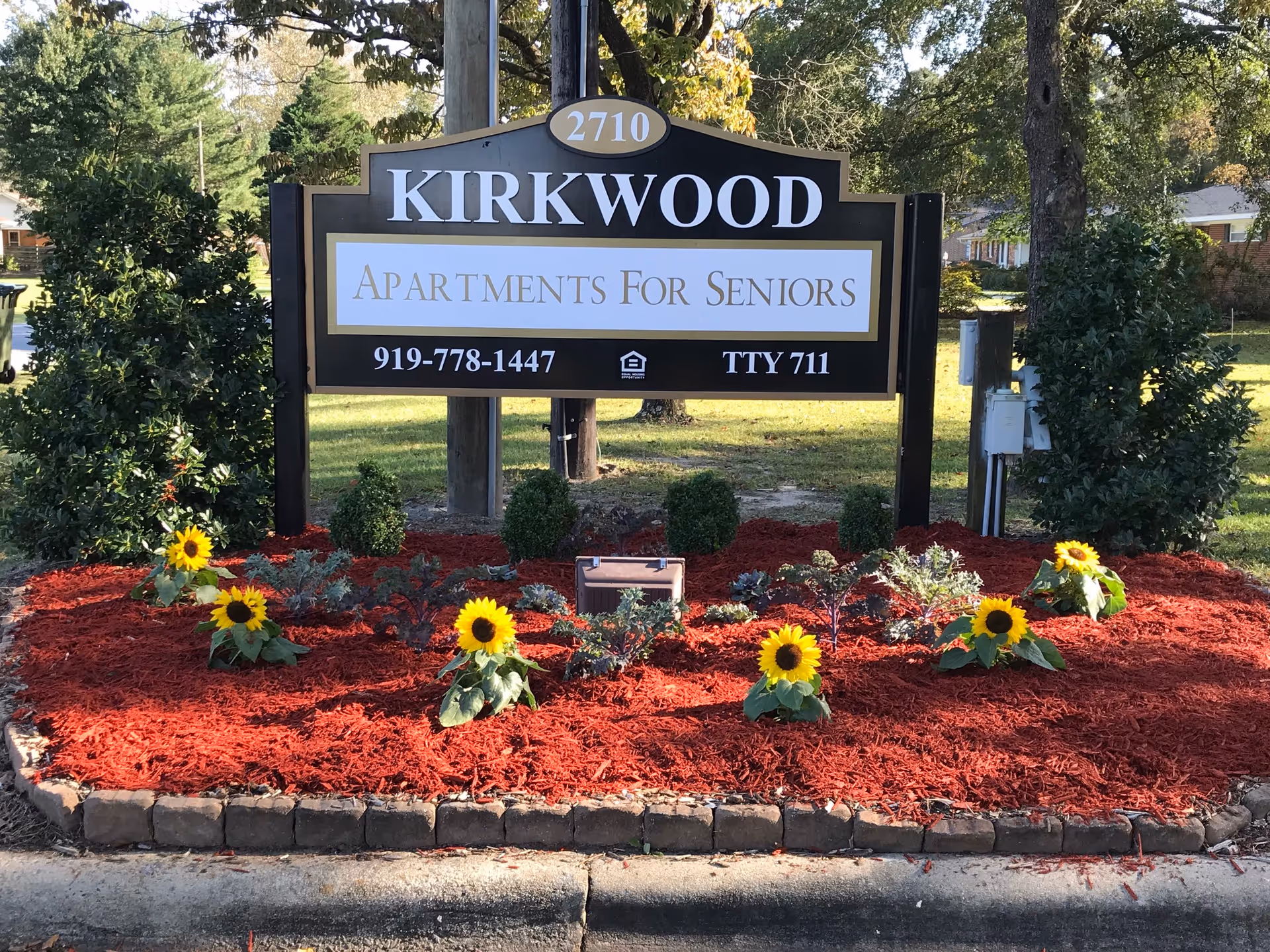 A landscaped entrance sign reading "KIRKWOOD Apartments For Seniors" with sunflowers and red mulch in front.