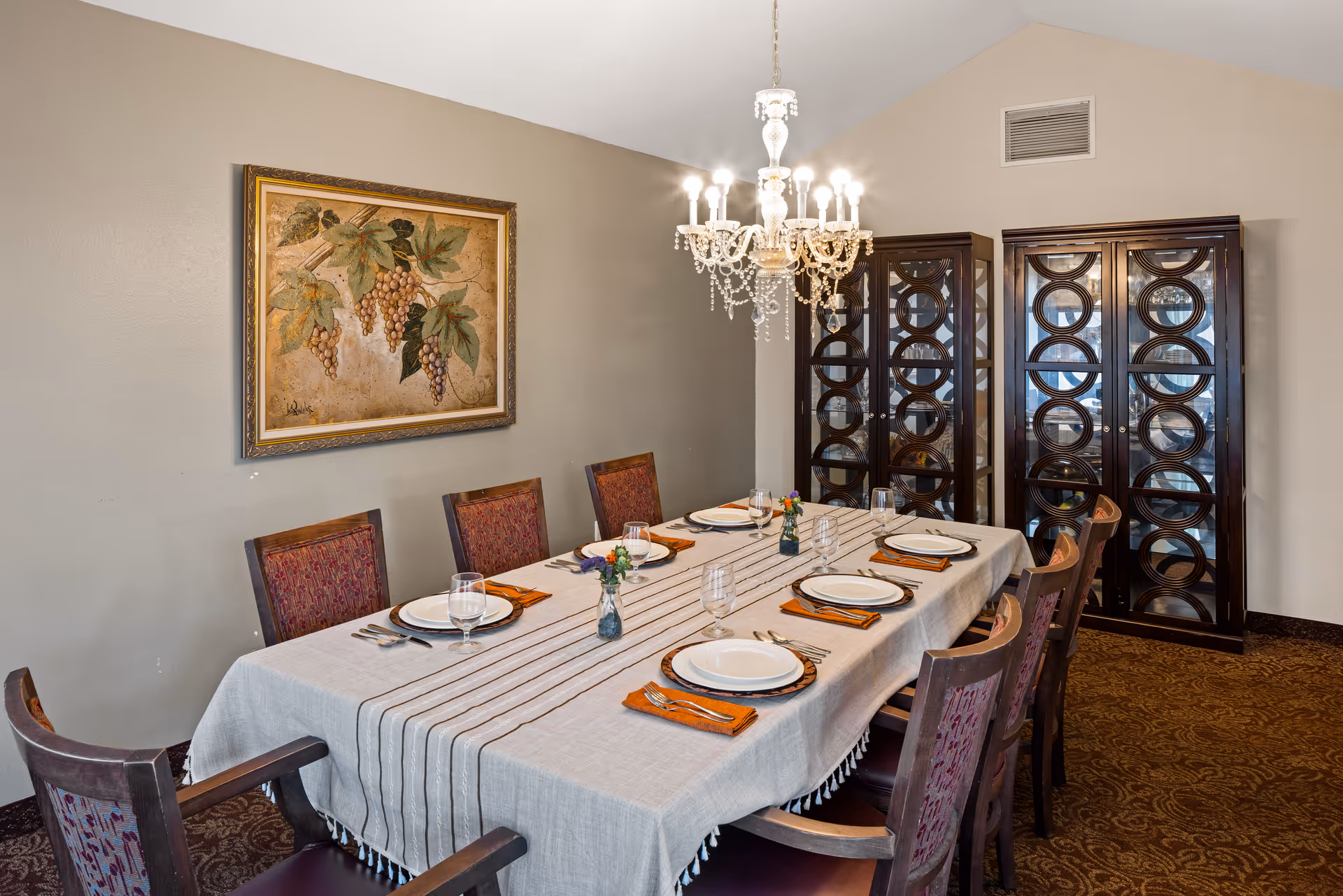 A formal dining room with a long rectangular table covered with a light-colored tablecloth and set for eight people with plates, glasses, and orange napkins. The room features a decorative chandelier hanging above the table, two dark wooden cabinets with glass doors in the background, and a framed painting of grapes on the wall.