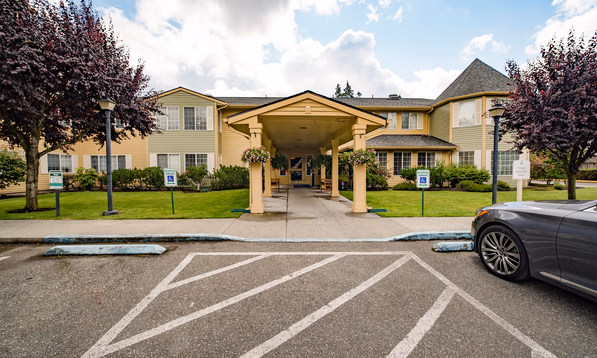 Front exterior view of Brookdale Everett facility showing a covered entrance with hanging flower baskets, two purple-leaved trees on either side, green lawn, and a parked gray car on the right side.