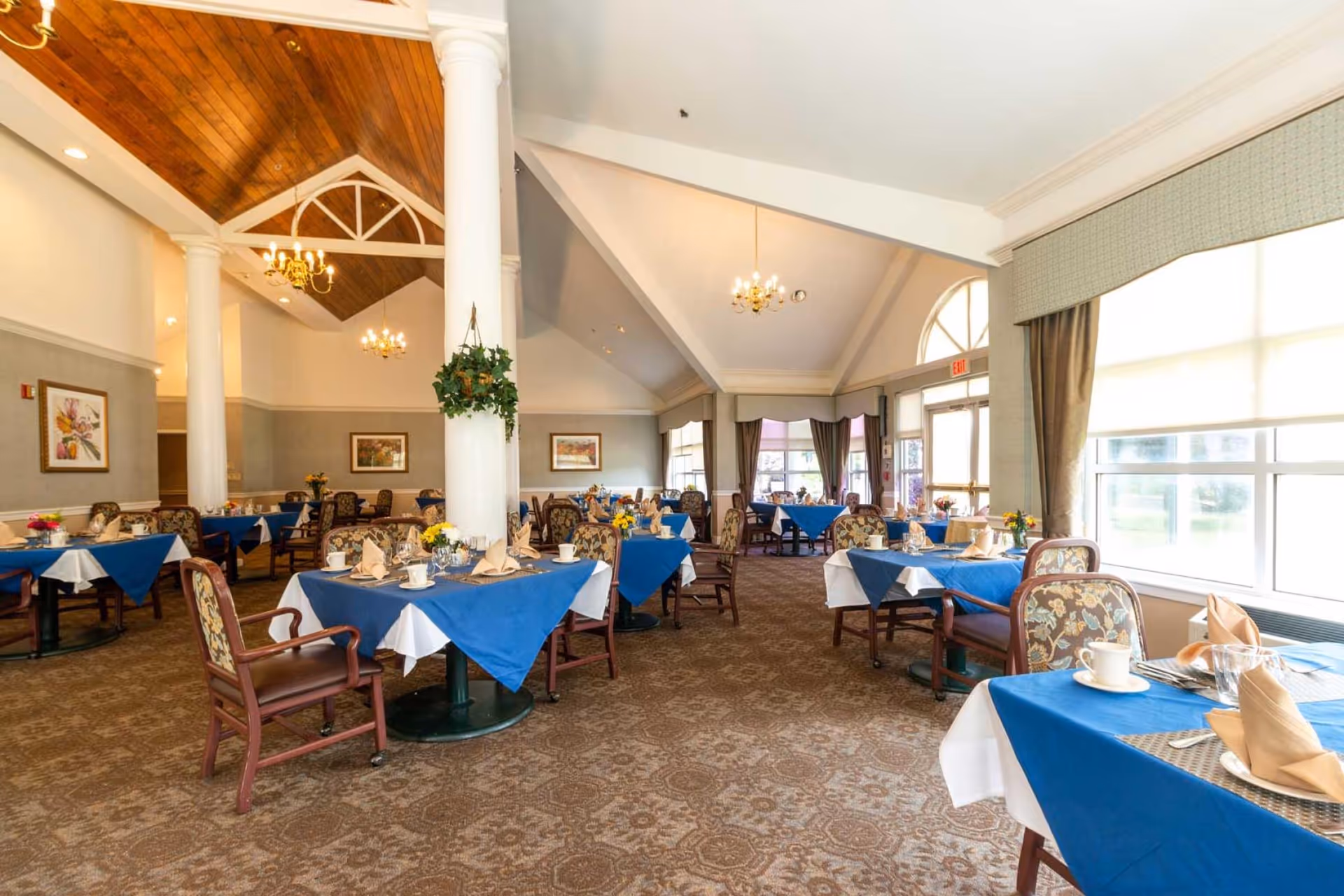 Spacious senior dining room with multiple round tables set with blue tablecloths, upholstered chairs, tall white columns, chandeliers, and large windows.