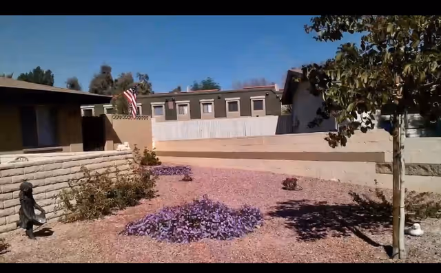 Outdoor view of a landscaped area at Beebe’s assisted living facility featuring a tree, purple flowering plants, a small statue, and buildings in the background under a clear blue sky.
