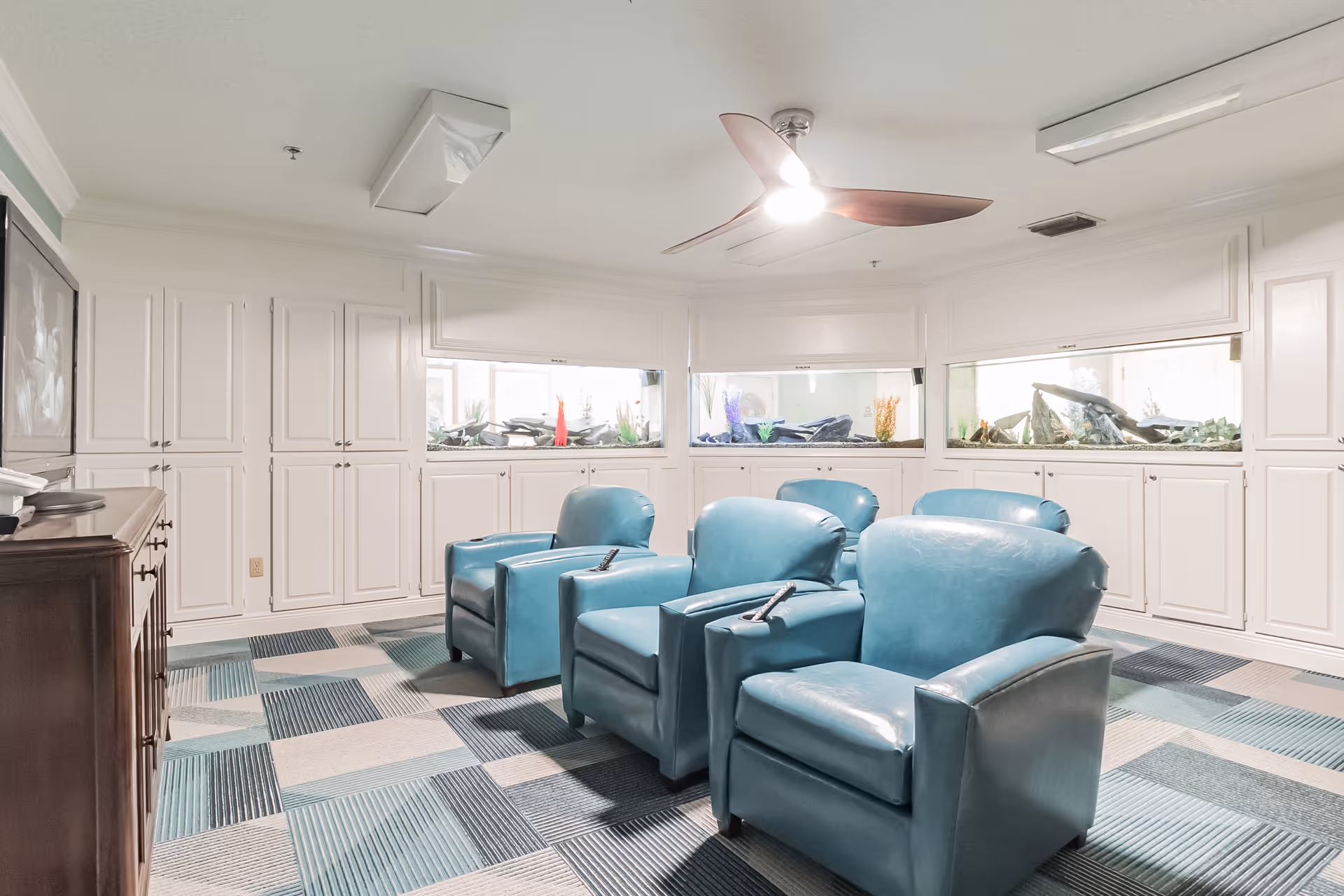 Bright common room with four teal leather recliner chairs facing a TV, built-in white cabinets and long aquariums along the wall.
