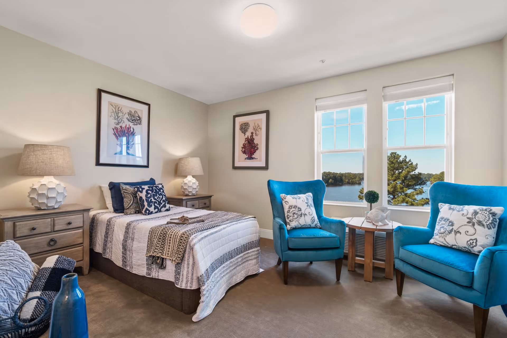 A cozy bedroom with a single bed covered in a striped quilt and decorative pillows. Two bedside tables with matching lamps flank the bed. Two bright blue armchairs with patterned cushions are positioned near two large windows that offer a view of a lake and trees outside. The walls are adorned with framed coral artwork, and a small wooden side table sits between the chairs.