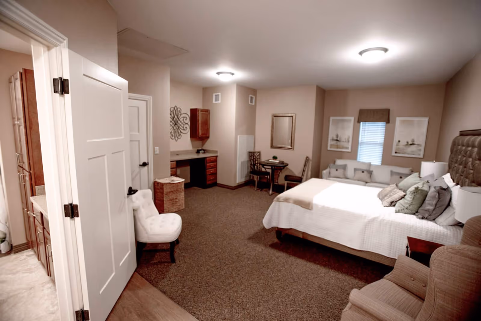 A spacious bedroom in a senior living facility featuring a large bed with multiple pillows, a small round table with two chairs, a white cushioned chair near the door, a beige armchair, and a sofa against the wall under two framed pictures and a window with blinds. The room has beige walls, carpeted floor, and ceiling lights. A partially open door reveals a bathroom with a shower and wooden cabinets.