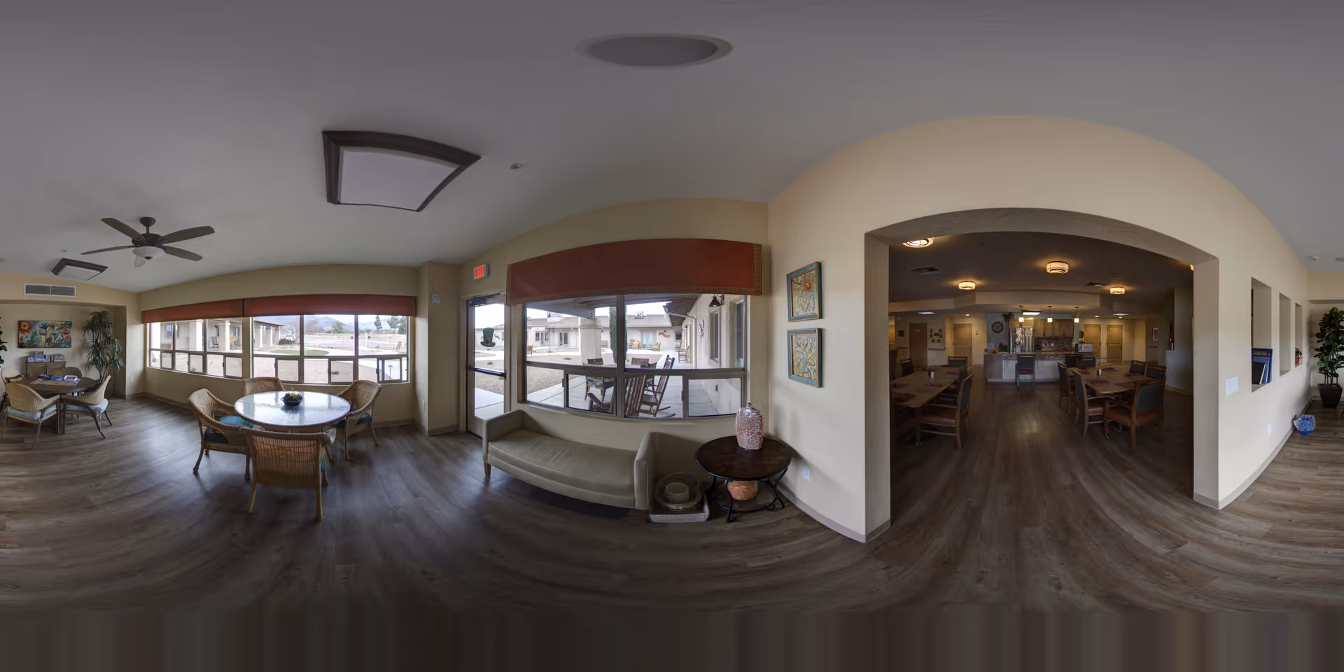 A spacious and well-lit common area in an assisted living facility featuring multiple seating arrangements including round tables with wicker chairs, a cushioned bench by the window, and a dining area with wooden tables and chairs visible through an arched doorway. Large windows provide natural light and views of an outdoor patio area. The room has wood flooring, ceiling fans, and neutral-colored walls with decorative artwork.