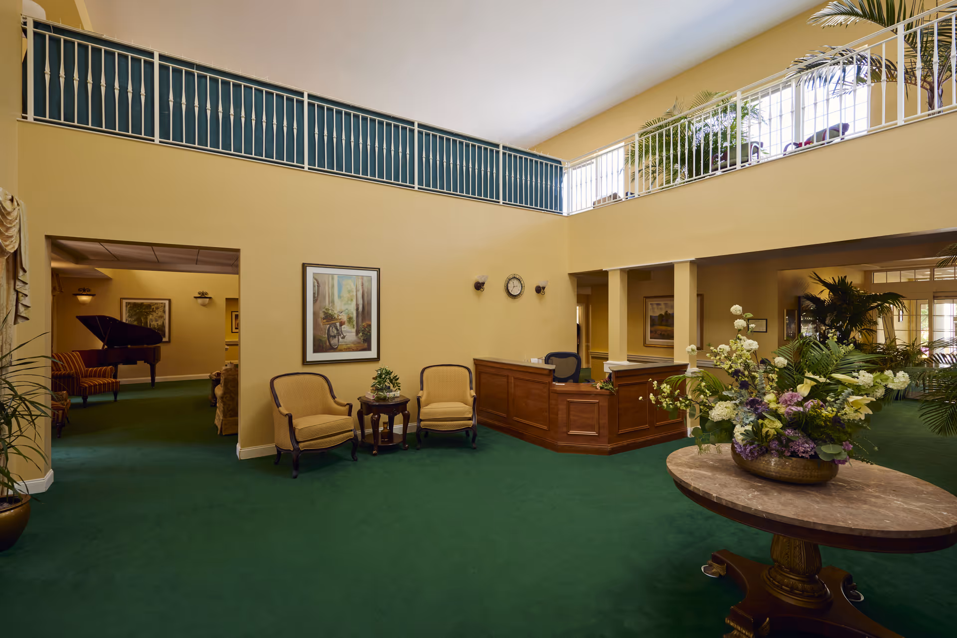 A spacious senior living facility lobby with green carpet, beige walls, and a wooden reception desk. Two upholstered chairs with a small round table are placed against the wall, which has a framed painting and a clock. A large round table with a floral arrangement is in the foreground. The area has a high ceiling with a balcony railing above and large windows letting in natural light. In the background, there is a room with a grand piano and additional seating.