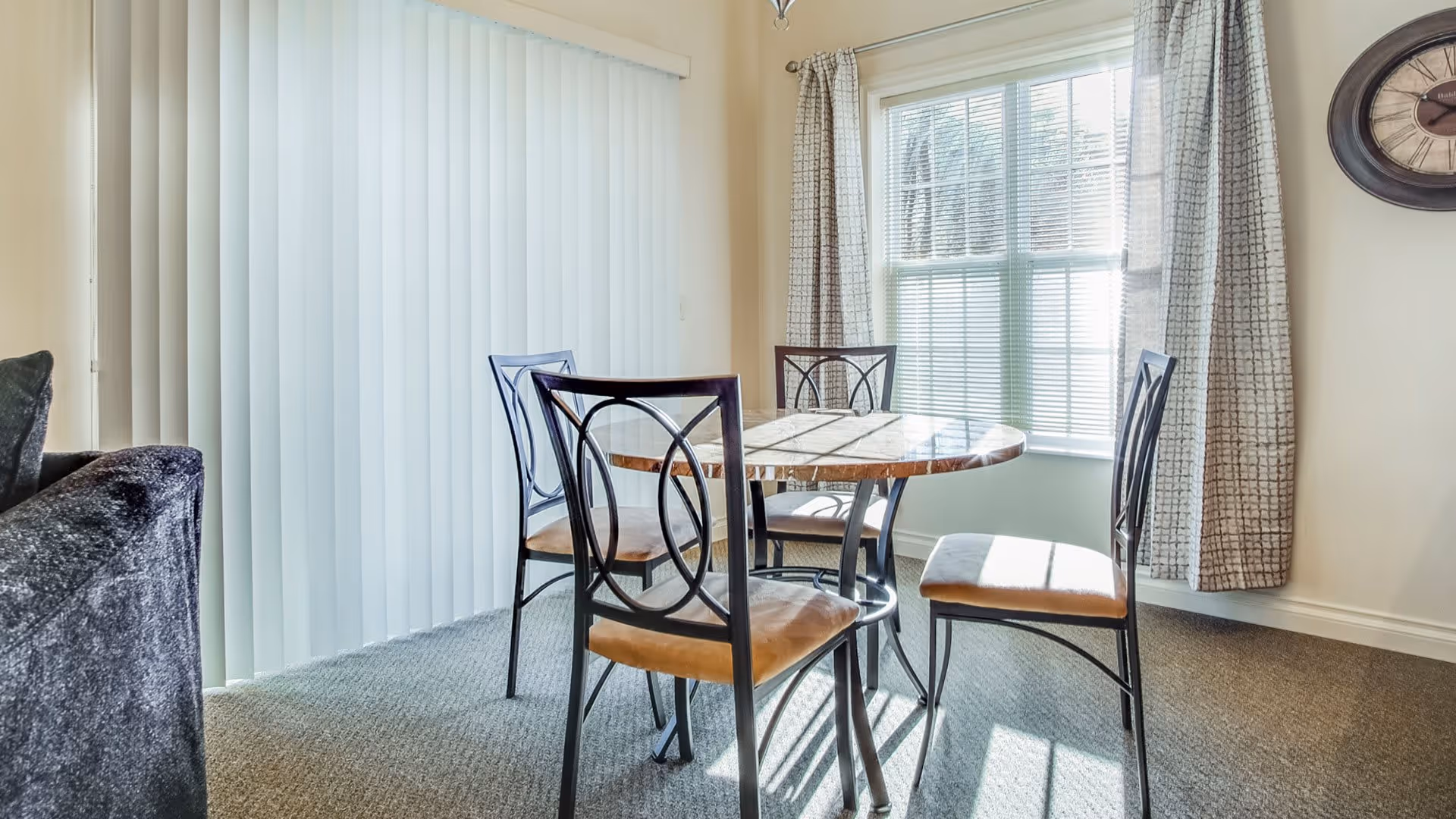 A bright dining area with a round wooden table and four metal chairs with cushioned seats. The room has beige walls, a large window with blinds and patterned curtains, and vertical blinds covering a sliding glass door. A wall clock is visible on the right side.