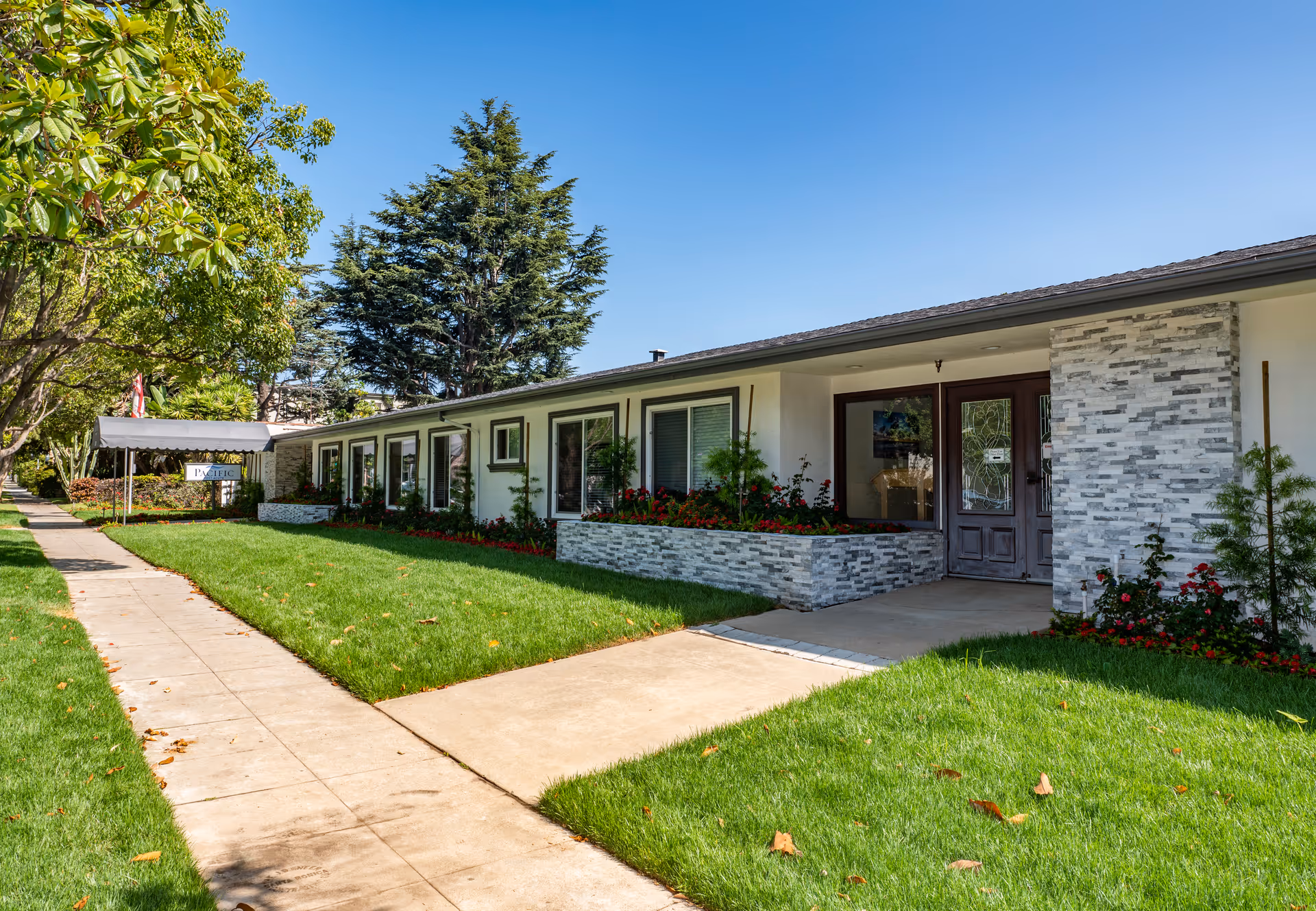 Exterior view of a single-story building with a well-maintained lawn and flower beds. The building has a stone facade around the entrance and multiple windows along the side. A sidewalk runs parallel to the building, and there are trees and shrubs along the walkway under a clear blue sky.