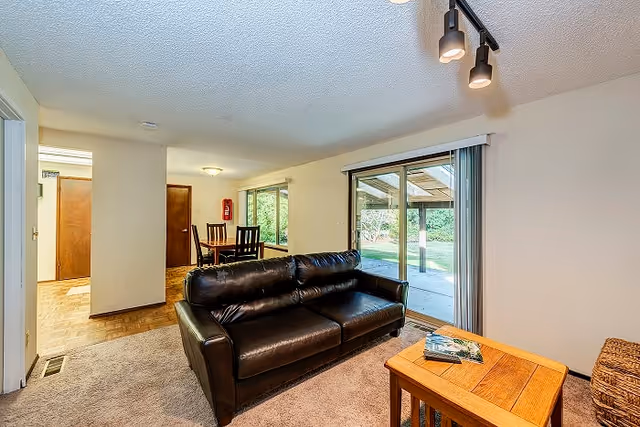 Interior view of a living room with a black leather sofa facing a wooden coffee table with magazines on it. Behind the sofa is a dining area with a table and chairs next to large windows. Sliding glass doors lead to a covered outdoor patio with greenery visible outside.