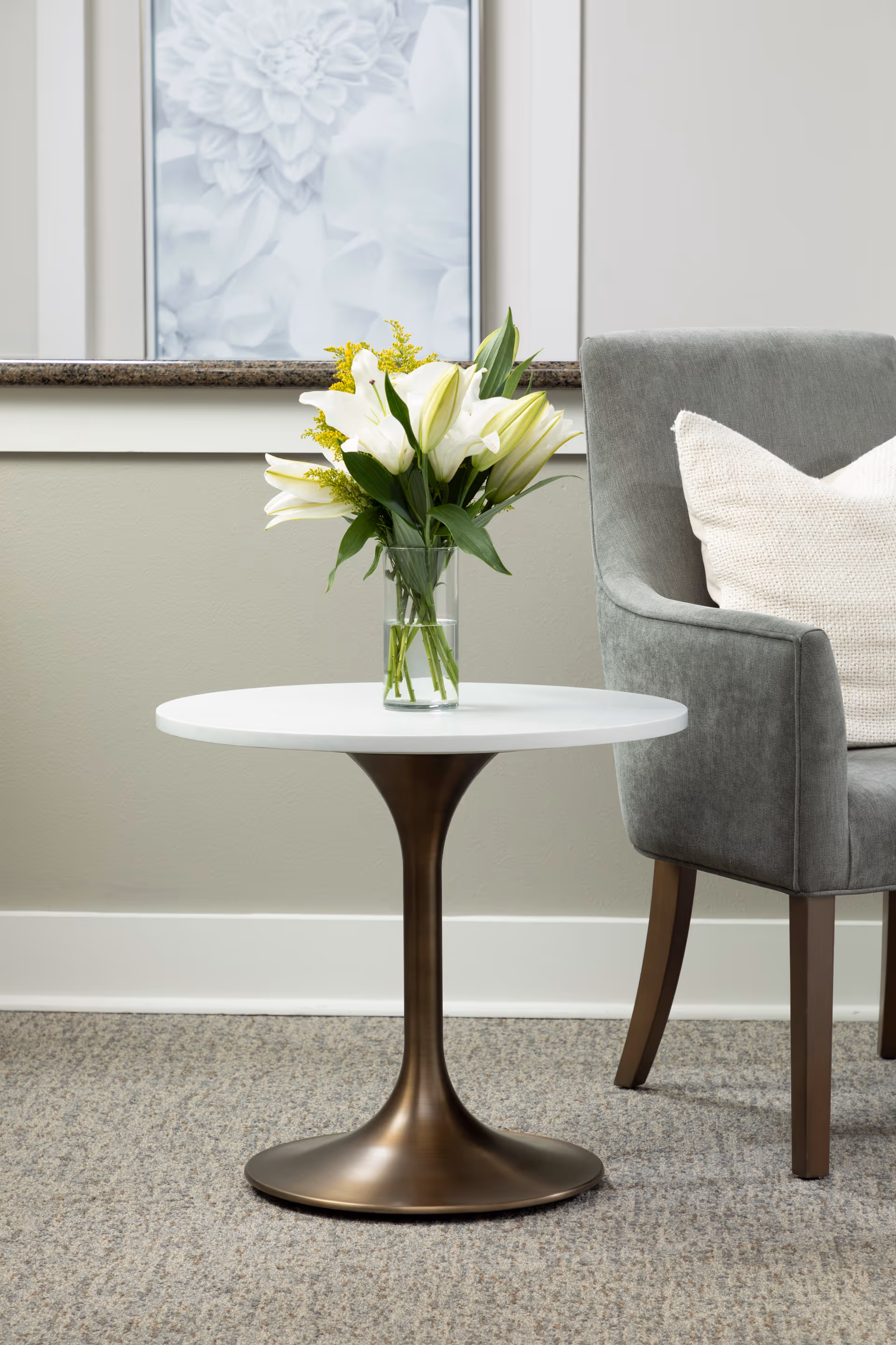 A modern round white table with a bronze pedestal base holding a clear glass vase filled with white lilies and yellow flowers, next to a gray upholstered chair with a white cushion, set against a beige wall with a framed floral artwork.
