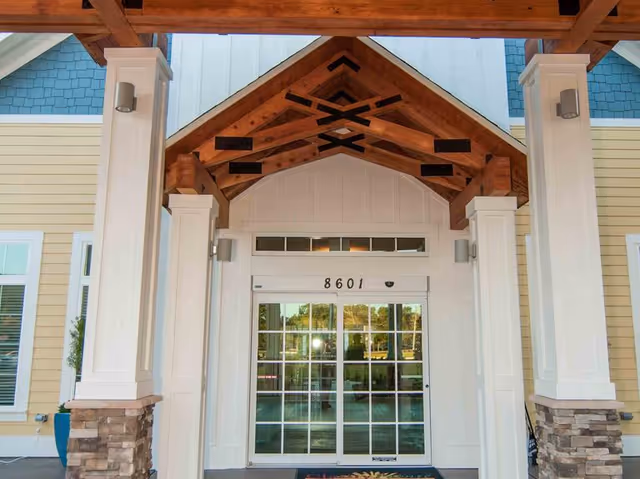 Entrance of a building with a covered porch featuring wooden beams and white columns with stone bases. The building has yellow siding and blue shingles near the roof. The glass double doors display the number 8601 above them.