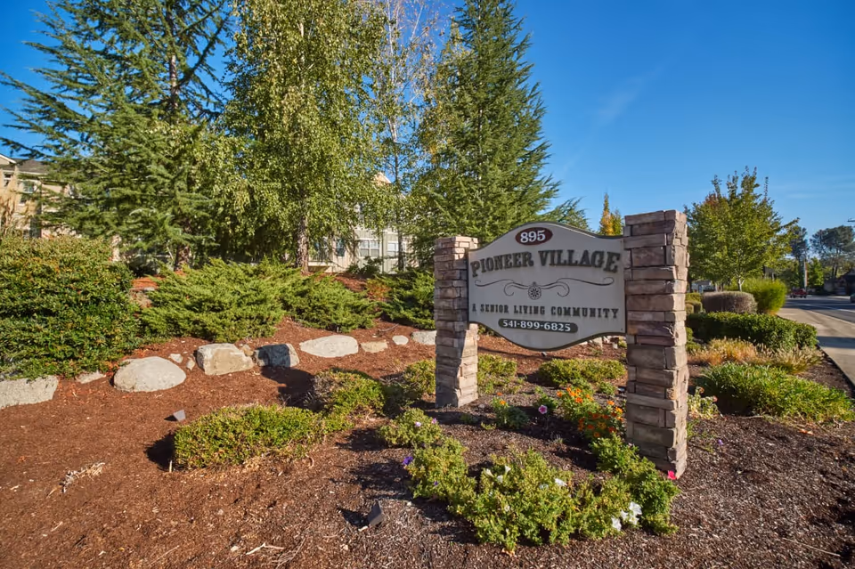 Stone entrance sign for Pioneer Village amid landscaped shrubs, rocks and trees under a blue sky.