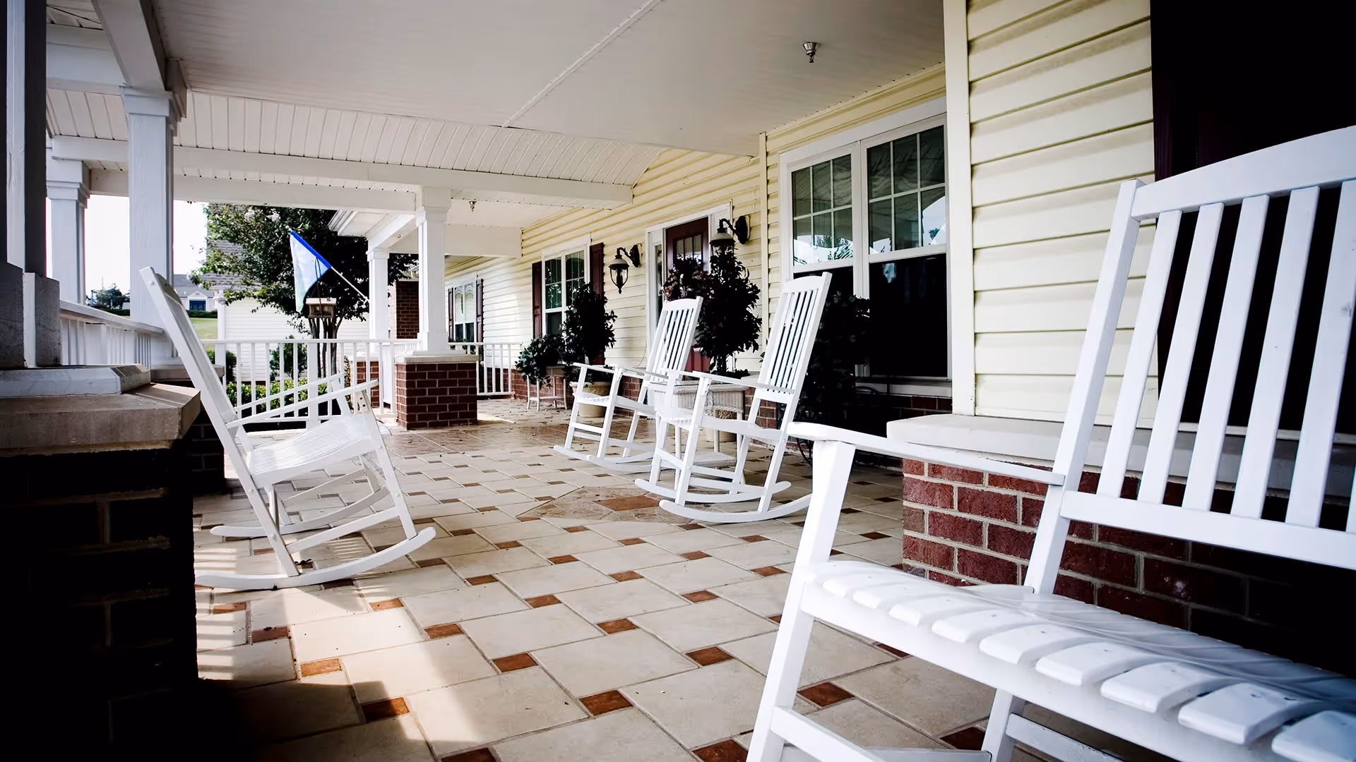 A covered porch area with several white wooden rocking chairs arranged along the wall of a building with yellow siding and large windows. The floor is tiled with a pattern of beige and brown squares. There are potted plants and outdoor lighting fixtures mounted on the wall.