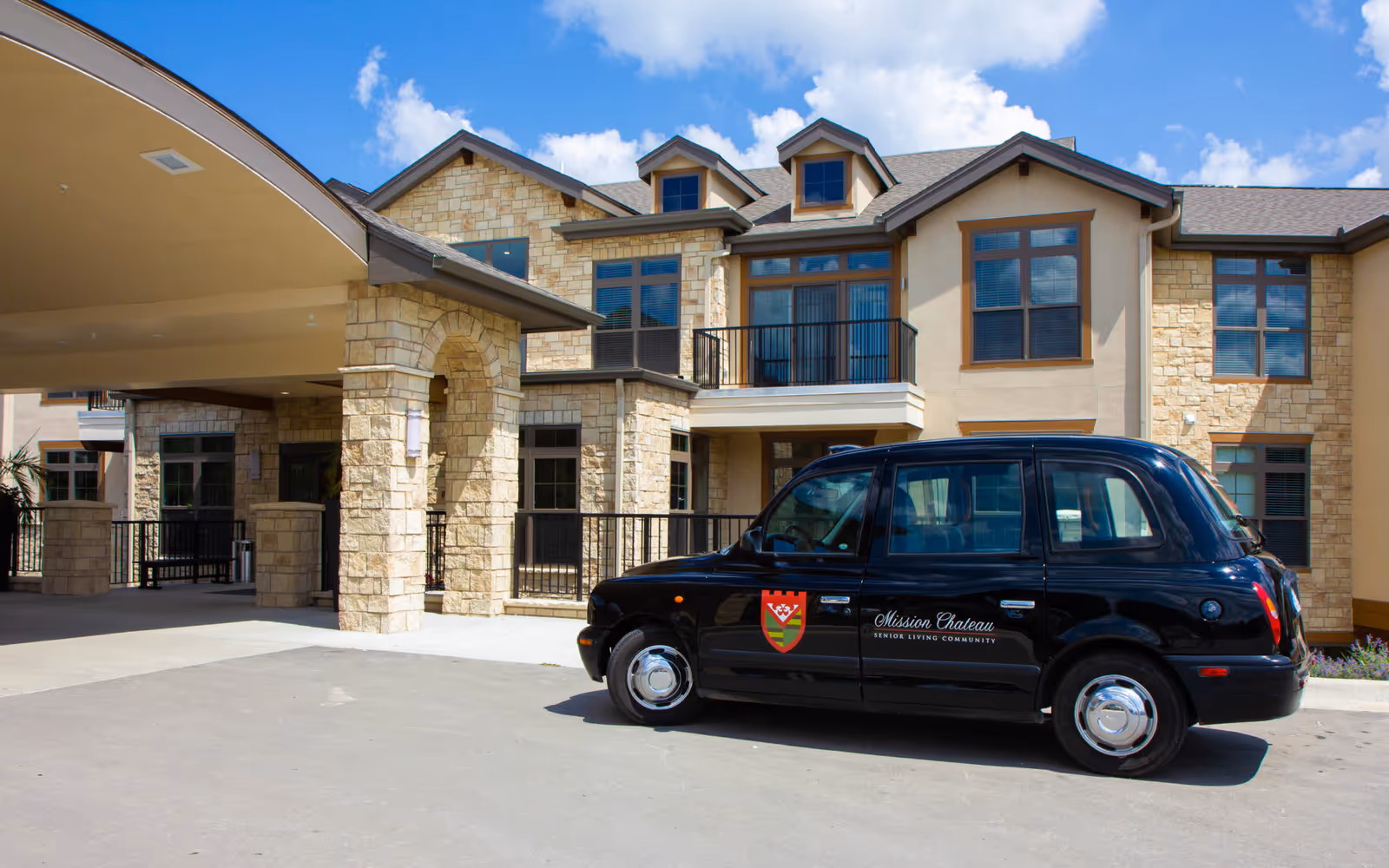 Exterior view of Mission Chateau Senior Living facility showing a stone and stucco building with multiple windows and a covered entrance. A black vehicle with the Mission Chateau Senior Living Community logo is parked in front of the building under a bright blue sky with scattered clouds.