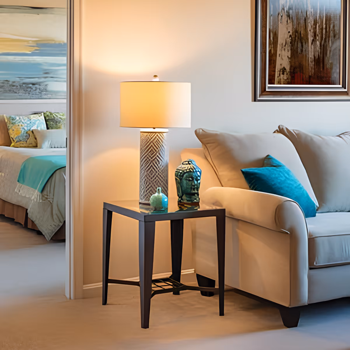 A cozy living room area with a beige sofa adorned with a teal cushion. Next to the sofa is a small dark wooden side table holding a decorative lamp with a patterned base and two small ornamental objects. In the background, a bedroom is partially visible with a bed covered in light bedding and teal accents, along with a large abstract painting on the wall.