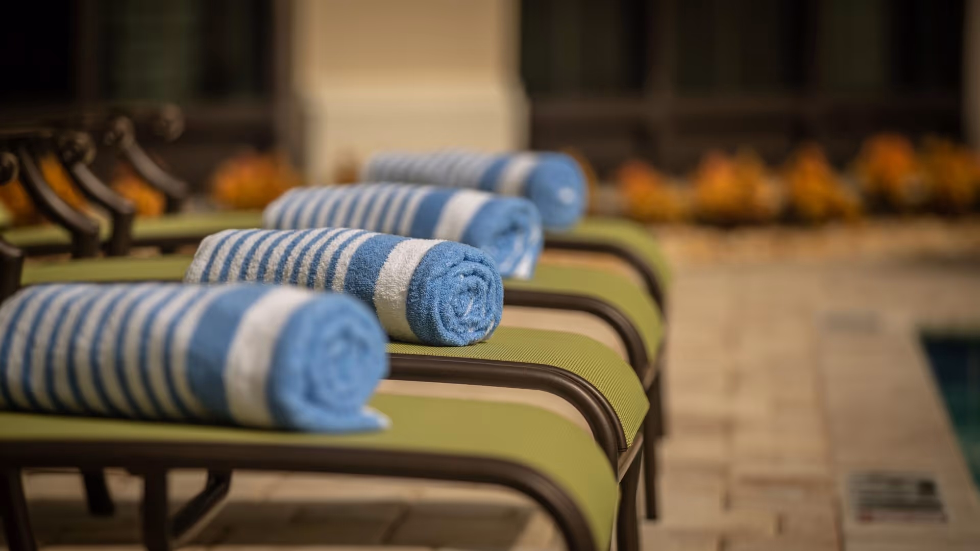 Close-up view of several green lounge chairs lined up outdoors, each with a neatly rolled blue and white striped towel placed on top, with a blurred background of a poolside area and some plants.