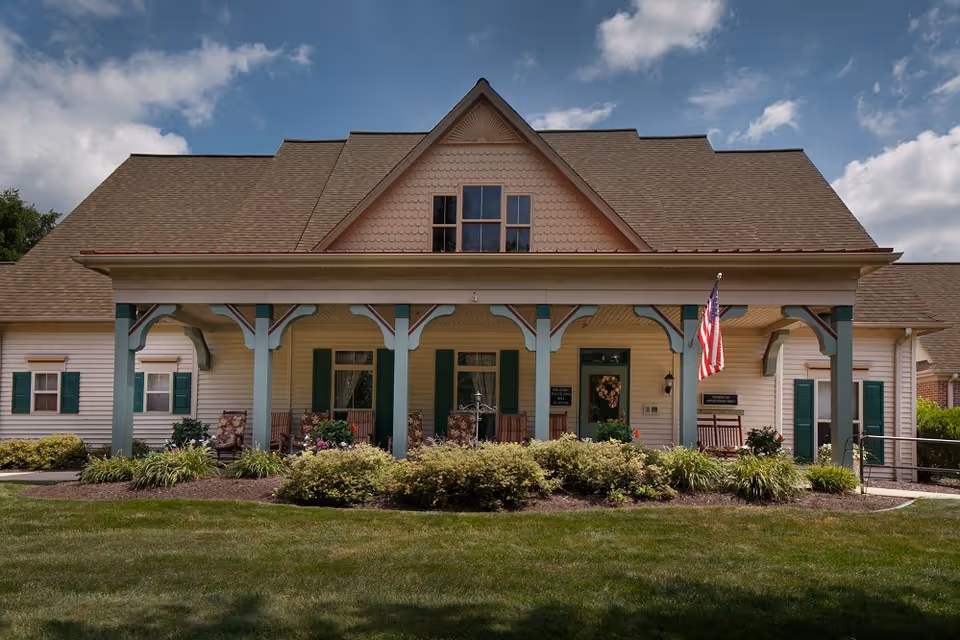Front facade of a cottage-style building with a covered porch, rocking chairs, landscaped shrubs, and an American flag.
