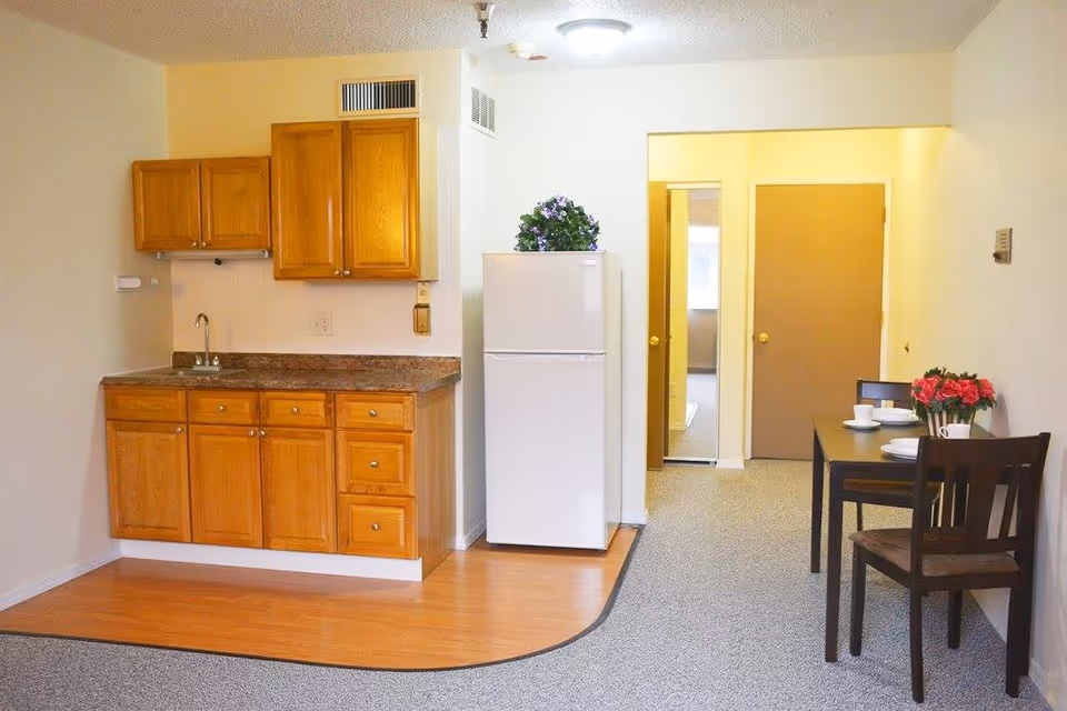 Interior view of a small kitchen and dining area in a retirement center. The kitchen has wooden cabinets, a countertop with a sink, and a white refrigerator with a plant on top. Adjacent to the kitchen is a small dining table set with two chairs, white dishes, and a vase with red flowers. The floor transitions from wood in the kitchen area to carpet in the dining and hallway areas. Two closed doors and an open doorway are visible in the background.