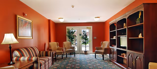 A cozy sitting room with red walls, patterned carpet, a striped sofa, several armchairs, and a wooden shelving/entertainment unit by glass doors.